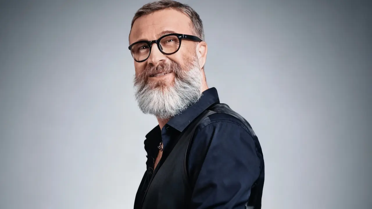 Un uomo con barba bianca e occhiali neri guarda sorridendo verso la fotocamera su sfondo neutro.