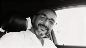 Un uomo con occhiali e barba corta sorride seduto all'interno di un'auto