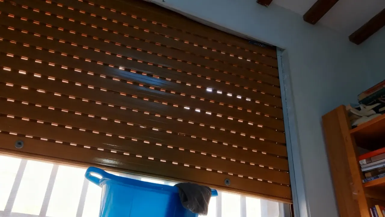 Tapparella marrone parzialmente abbassata vista dall'interno con secchio blu nelle vicinanze