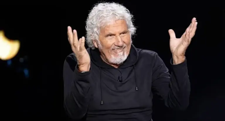 Un uomo con capelli ricci bianchi e barba sorride mentre gesticola con le mani.