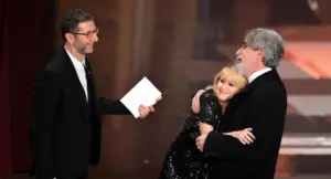 Tre persone sul palco, una delle quali abbraccia affettuosamente un uomo anziano mentre un'altra osserva sorridendo