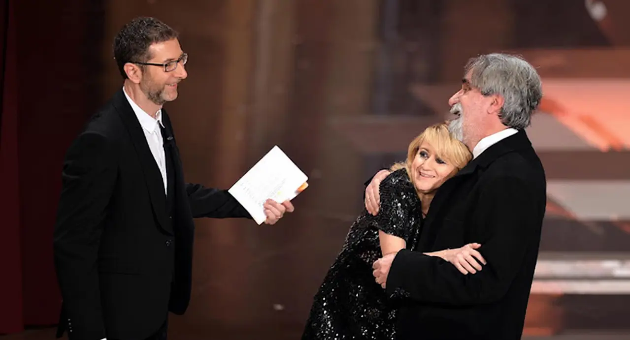 Tre persone sul palco, una delle quali abbraccia affettuosamente un uomo anziano mentre un'altra osserva sorridendo